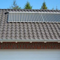 Auf dem Dach sind glänzende Solarpanels installiert, die sich harmonisch in die wellenförmigen Ziegel einfügen, umgeben von Natur.