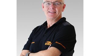 Ein lächelnder Mann in einem schwarzen Poloshirt mit einem Logo, der selbstbewusst mit verschränkten Armen posiert.