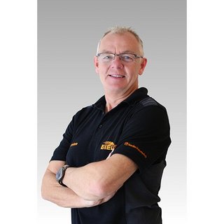 Ein lächelnder Mann in einem schwarzen Poloshirt mit einem Logo, der selbstbewusst mit verschränkten Armen posiert.