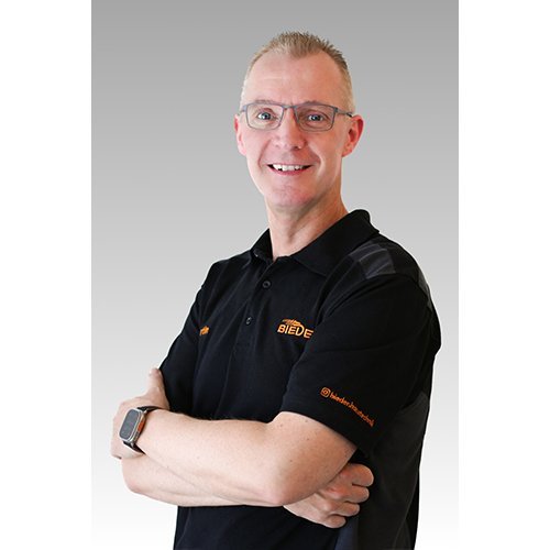 Der Mann trägt ein schwarzes Poloshirt mit dem Logo 'BIEDE' und hat eine freundliche Ausstrahlung, während er in die Kamera schaut.