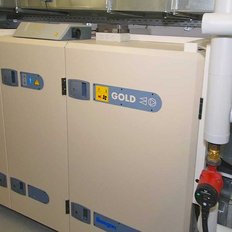 Drei große, beige Geräte mit blauen und grauen Aufklebern stehen in einem modernen Raum, umgeben von Rohren und technischen Anlagen.