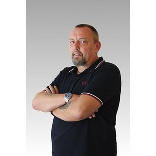 Mit einem selbstbewussten Blick steht der Mann mit tätowierten Armen und einem Ohrstecker vor einem neutralen Hintergrund.