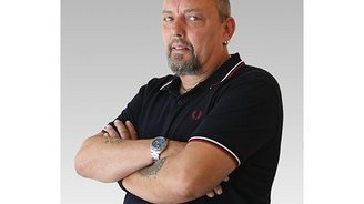 Mit einem selbstbewussten Blick steht der Mann mit tätowierten Armen und einem Ohrstecker vor einem neutralen Hintergrund.