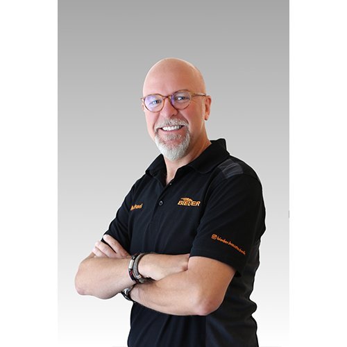 Der Mann mit Glatze und Bart strahlt Freundlichkeit aus, während er in einem schwarzen Shirt mit Logo posiert.