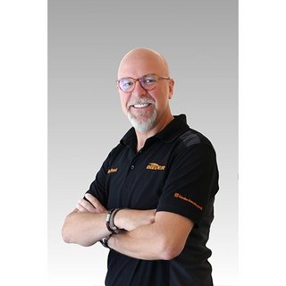 Der Mann mit Glatze und Bart strahlt Freundlichkeit aus, während er in einem schwarzen Shirt mit Logo posiert.