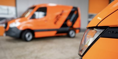 Ein orangefarbener Transporter mit einem klaren Scheinwerfer ist im Vordergrund, während ein weiterer im Hintergrund unscharf ist.