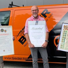 Die Männer zeigen ihre Meisterbriefe, während der orangefarbene Firmenwagen im Hintergrund sichtbar ist.