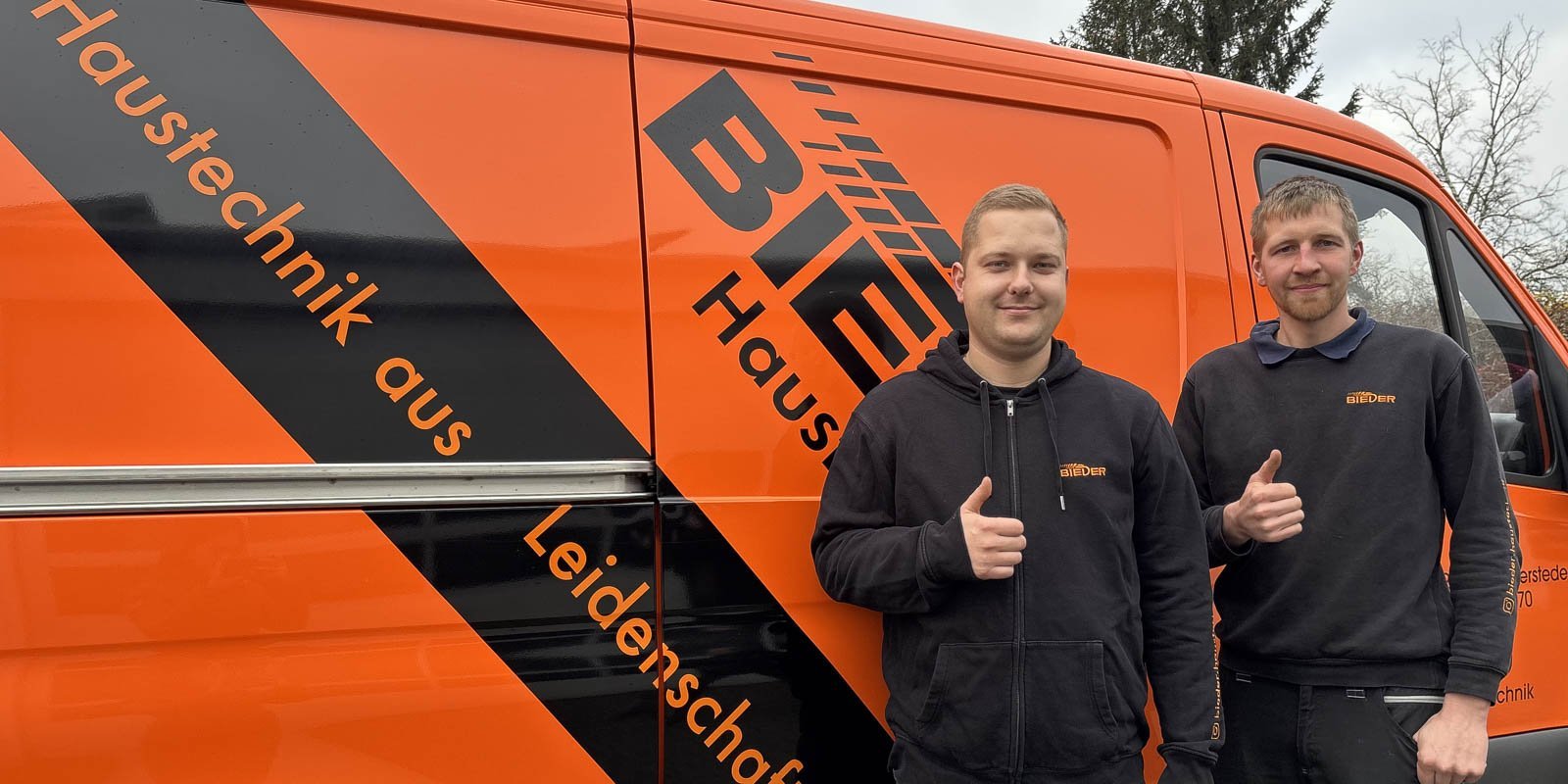 Zwei Männer stehen vor einem orangefarbenen Lieferwagen mit dem Schriftzug 'BIEDER Haustechnik aus Leidenschaft'.