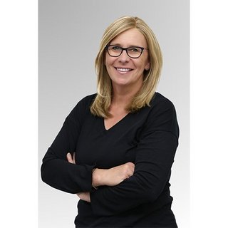 Eine lächelnde Frau mit blonden Haaren und Brille steht mit verschränkten Armen vor einem hellen Hintergrund.