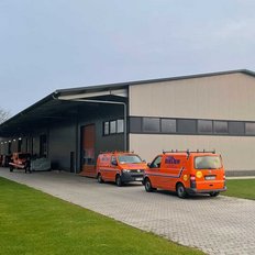 Das Bild zeigt ein Lagerhaus mit einem überdachten Bereich, umgeben von Bäumen und mehreren orangefarbenen Fahrzeugen.