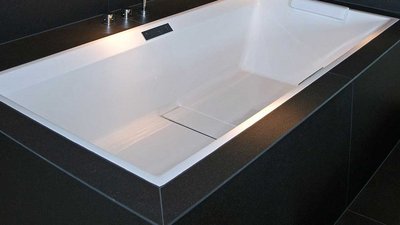 In einem modernen Badezimmer befindet sich eine schlichte, weiße Badewanne, umgeben von dunklen Oberflächen und sanftem Licht.