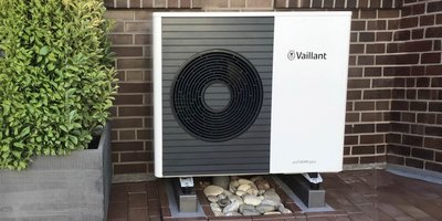 Energieeffiziente Heiztechnik in einem stilvollen Außenbereich Eine moderne Wärmepumpe steht neben einem großen, grünen Pflanzenbehälter auf einem gepflasterten Boden.