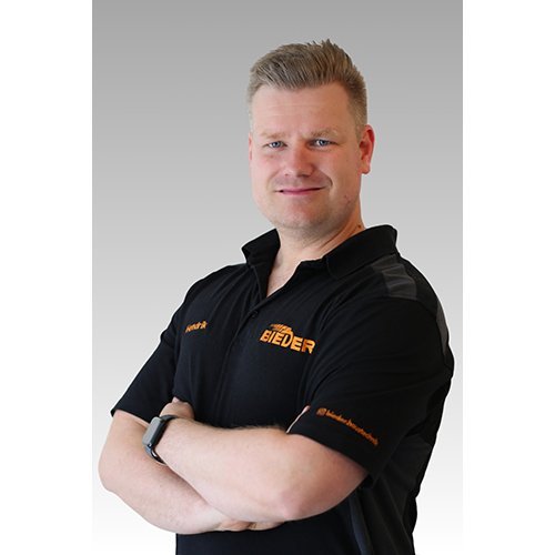 In einem schwarzen Shirt mit dem Schriftzug 'BIEDER' präsentiert sich ein lächelnder Mann, der eine positive Ausstrahlung hat.