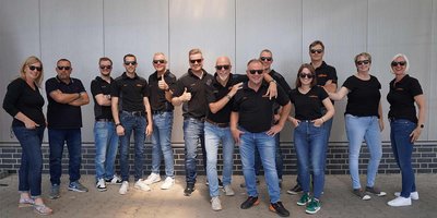 Die Gruppe besteht aus Männern und Frauen unterschiedlichen Alters, die in lässiger Kleidung und Sonnenbrillen posieren.