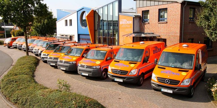 Vor einem stilvollen, blauen und orangefarbenen Gebäude stehen mehrere orangefarbene Lieferwagen, die für eine Firma namens BIEDER werben.