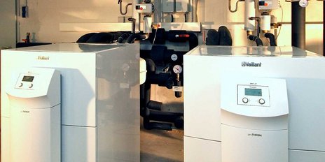 In einem technischen Raum sind zwei Vaillant-Heizsysteme sichtbar, die mit verschiedenen Rohren und Messgeräten verbunden sind.