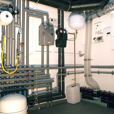 In einem technischen Raum sind zahlreiche Rohre und Geräte sichtbar, die für die Heizungs- und Wasserversorgung zuständig sind.