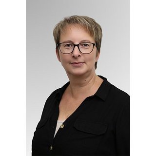 Eine Frau mit kurzen, blonden Haaren und Brille trägt ein schwarzes Hemd und schaut direkt in die Kamera.